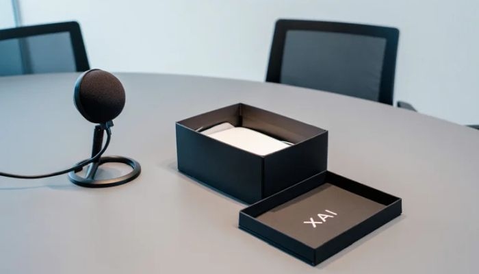 Symbolic picture showing a podcastsituation with office table an two chairs. On the table a microphone and a opened black-box. The cover of the box shows the letters XAI inside.