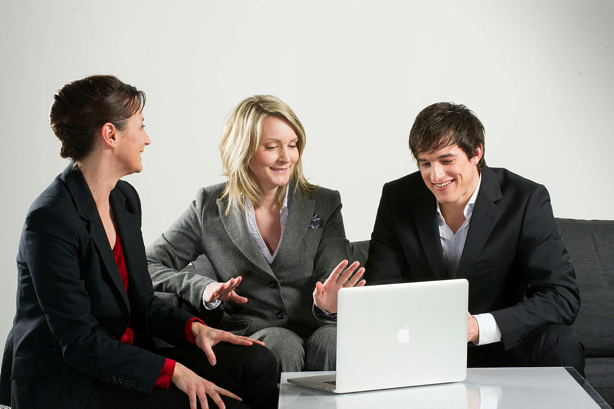 Drei Personen sitzen um einen Schreibtisch und schauen auf einen Laptop und besprechen sich