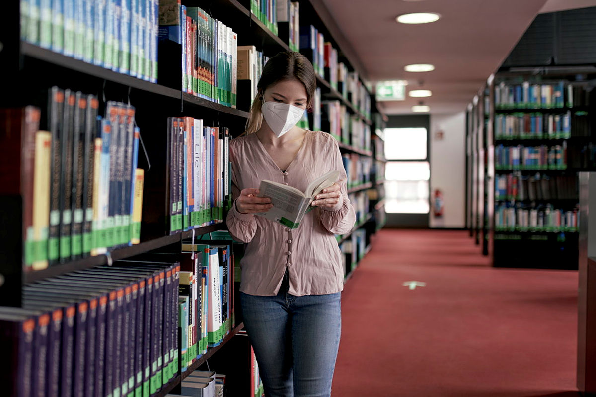 Eine WIssenschaftlerin steht in der Bibliothek an ein Regal gelehnt